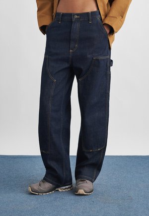 Person iført løse mørkeblå denim cargobukser med kontrastsyninger og grå sneakers, stående på blåt tæppe.