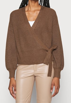 Femme portant un pull en maille brun avec un détail de nouage sur le côté et un pantalon en cuir synthétique beige, debout les mains détendues le long du corps.