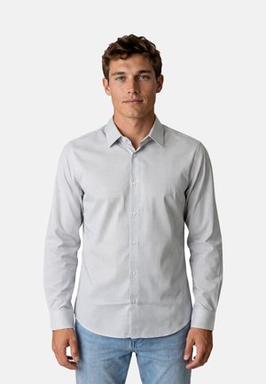 Giovane uomo in piedi di fronte, indossa una camicia a maniche lunghe grigia chiara a fantasia con bottoni e jeans blu su sfondo bianco.