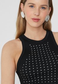 Czarna, ryflowa tank top z okrągłym dekoltem i ozdobami z nitów ułożonymi w pionowe linie. Modelka nosi duże, białe, owalne kolczyki.