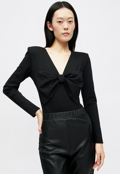 Μαύρο κορμάκι με μακριά μανίκια και βαθύ V-ανοιγμένο λαιμό, που διαθέτει έντονη λεπτομέρεια φιόγκου μπροστά, συνδυασμένο με μαύρο παντελόνι με ψηλή μέση.