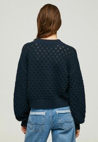 Granatowy dziergany sweter z teksturowanym wzorem diamentowym, o luźnym kroju, ze ściągaczami na rękawach i dole; noszony z jasnoniebieskimi jeansami.