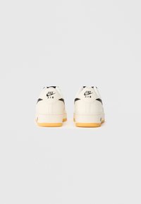 Tênis brancos de couro com detalhes pretos, apresentando uma sola de borracha e a marca "Nike Air" no calcanhar. Bico arredondado e colarinho acolchoado.