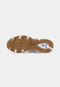 Semelle extérieure en caoutchouc brun et blanc d'une chaussure New Balance avec une bande de roulement à motifs et des éléments d’amorti bleus sur fond blanc.