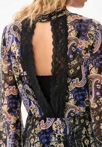 Robe longue à motifs avec une bordure en dentelle noire. Dotée d'un dos en V profond et de manches longues. Le tissu présente des motifs floraux et cachemire en bleu et crème.