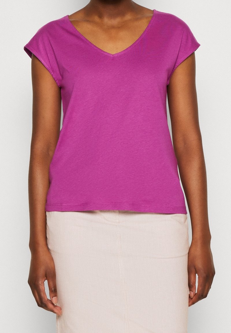 Femme portant un haut violet vif à manches et col en V et une jupe beige clair, visible des épaules jusqu'au milieu des cuisses sur un fond uni.