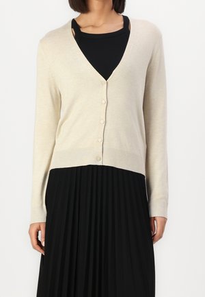Un mannequin porte un cardigan crème boutonné à col en V sur un haut noir et une longue jupe noire plissée, sur un fond blanc uni.