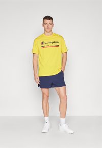 Camiseta de algodón amarilla con el logo "Champion New York" en negro, rojo y amarillo. Llevada con pantalones cortos azul marino y zapatillas blancas.