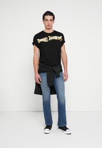 Μαύρο Τ-shirt με χρυσό γραφικό κείμενο, συνδυασμένο με μπλε τζιν παντελόνι και μαύρα αθλητικά παπούτσια. Το T-shirt έχει μανίκια που είναι γυρισμένα προς τα πάνω· το τζιν είναι εφαρμοστό.