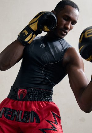 Boxer in black and gold gloves and black sleeveless shirt with red shorts, holding guard up in a fighting stance against a plain wall.