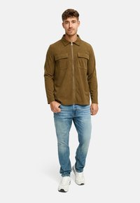 Mand iført en brun lynlås cordurojacket, lyseblå jeans og hvide sneakers, stående op ad en ensfarvet lys baggrund.