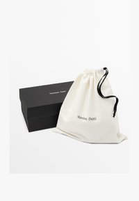 Een trekkoordtas van crème katoen met "Massimo Dutti" in zwart gedrukt, vergezeld van een zwarte rechthoekige doos.