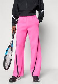 Φουξια αθλητικό παντελόνι με φαρδύ πόδι και μαύρες λωρίδες στο πλάι, κατασκευασμένο από μαλακό ύφασμα. Φορεμένο με μαύρο ζακέτα με φερμουάρ, το άτομο κρατάει ρακέτα τένις.