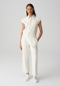 Combinaison en denim blanc avec des manches courtes, boutons sur le devant et poche poitrine. Associée à des jambes droites amples et des baskets blanches.