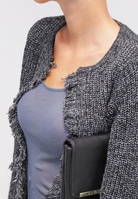 Svart och vit stickad cardigan med texturerade fransade kanter, bärs över en blå topp och håller en svart kuvertväska.