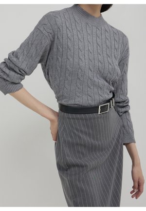 Femme portant un pull torsadé gris, une jupe grise à rayures fines et une ceinture noire, debout avec une main sur la hanche devant un fond uni.