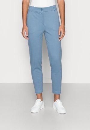 Personne portant un pantalon slim bleu clair avec fermeture par bouton et zip, associé à des baskets blanches et un haut blanc.