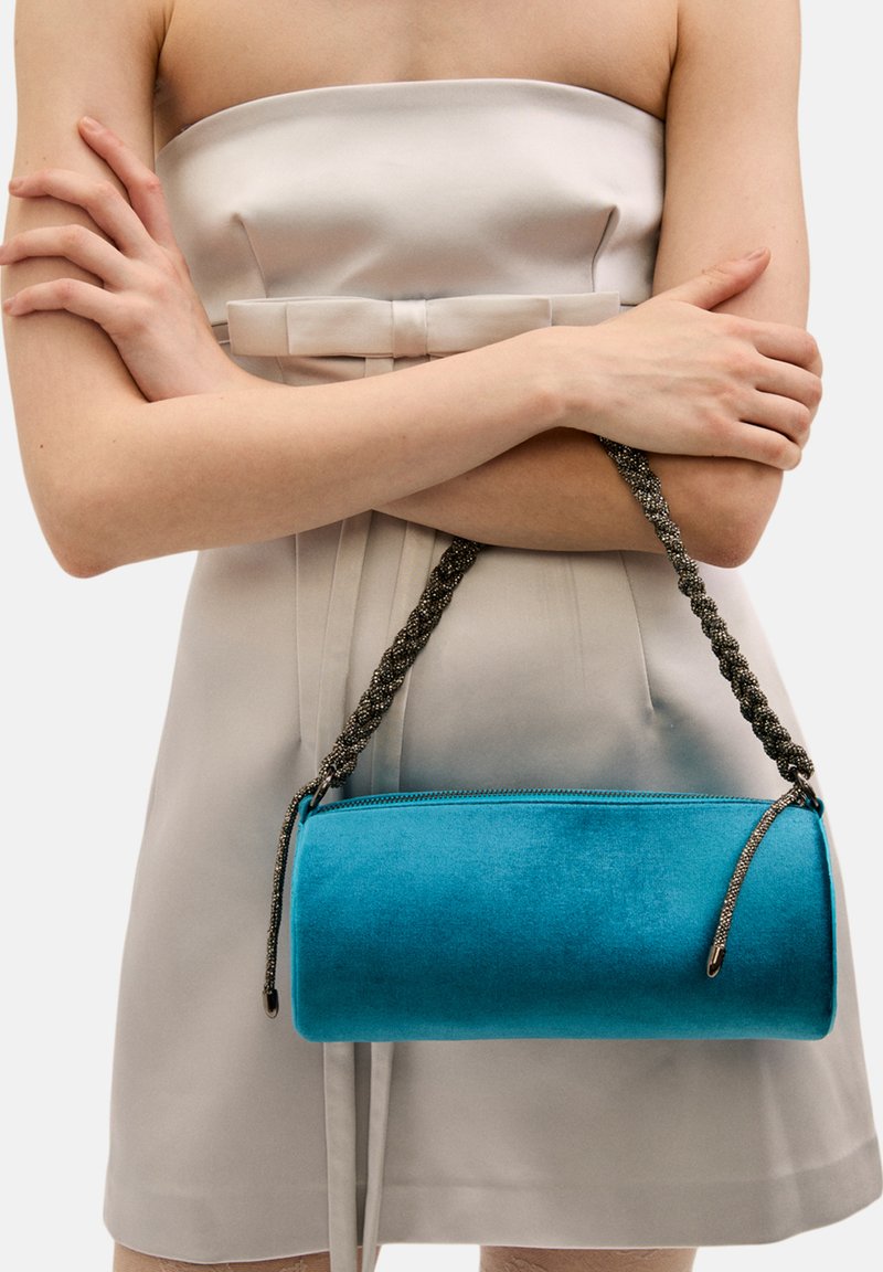 Turkos sammetshandväska med cylindrisk form, flätad metallrem och dragkedjestängning. Slät textur och livlig färg.