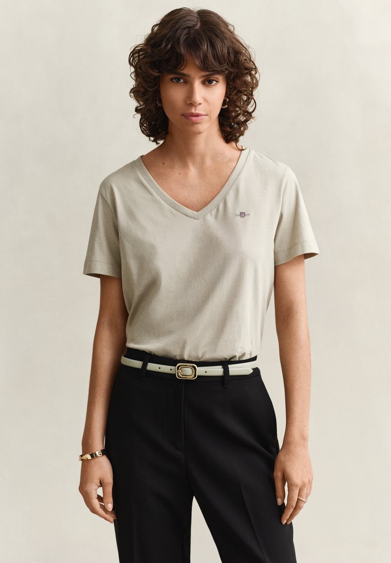Frau mit lockigem Haar trägt beige V-Ausschnitt T-Shirt, in schwarze Hose mit weißem Gürtel gesteckt, steht vor hellem einfarbigem Hintergrund.
