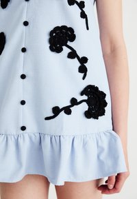 Blue dress with black floral embroidery, button details down the front, and a ruffled hem. Fabric appears textured and lightweight.