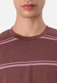 Brown cotton T-shirt with a round neckline, featuring two horizontal light pink stripes across the chest. Soft texture, regular fit.