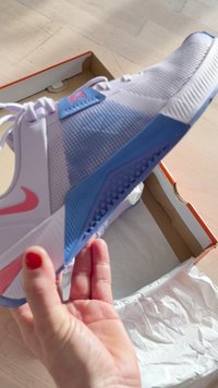 Hand hält einen weißen und blauen Sportschuh mit pinkem Nike-Logo über einem offenen Schuhkarton auf einem Holzfußboden.