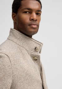 Beige houndstooth jas met een hoge kraag, voorzien van twee grote knopen en een gestructureerde stof, getoond op een model.