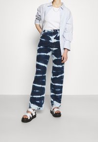 Blå tie-dye jeans med lös passform och vida ben. Tyget är denim med vita virvlande mönster, kombinerat med vita sandaler med remmar.