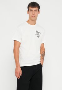 Tricou alb din bumbac cu guler rotund și mâneci scurte. Prezintă text imprimat negru pe pieptul stâng. Purtați cu pantaloni negri.