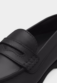 Mocassin en cuir noir avec des détails cousus et une sangle sur le dessus, présentant un design subtilement découpé.