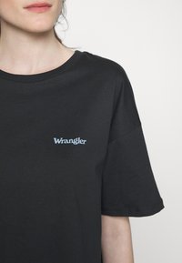 Femme portant un t-shirt noir à manches courtes avec un logo "Wrangler" bleu clair sur la poitrine gauche, sur fond blanc.