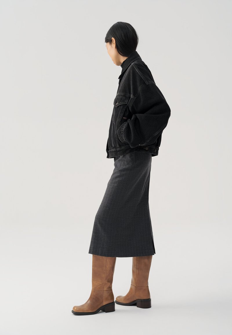 Giacca di jeans nera, gonna a righe grigie, stivali alti fino al ginocchio in pelle marrone con punta quadrata, su uno sfondo neutro.