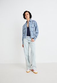 Chemise à carreaux bleue boutonnée portée sur un haut bleu marine, associée à un jean évasé clair et des baskets blanches avec des accents rouges.