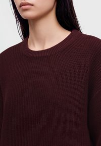 Femme portant un pull en maille bordeaux foncé à col rond, laissant apparaître son cou, la ligne de sa mâchoire et une partie de ses cheveux foncés.