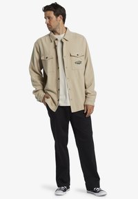 Beige fleece button-up shirt with two chest pockets and embroidered logo, paired with black pants and black sneakers.