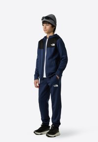 Marineblå treningsdress med svart øvre panel, laget av glatt stoff. Hvit glidelåslukking, logoaccenter, kombinert med svarte treningssko og grå lue.