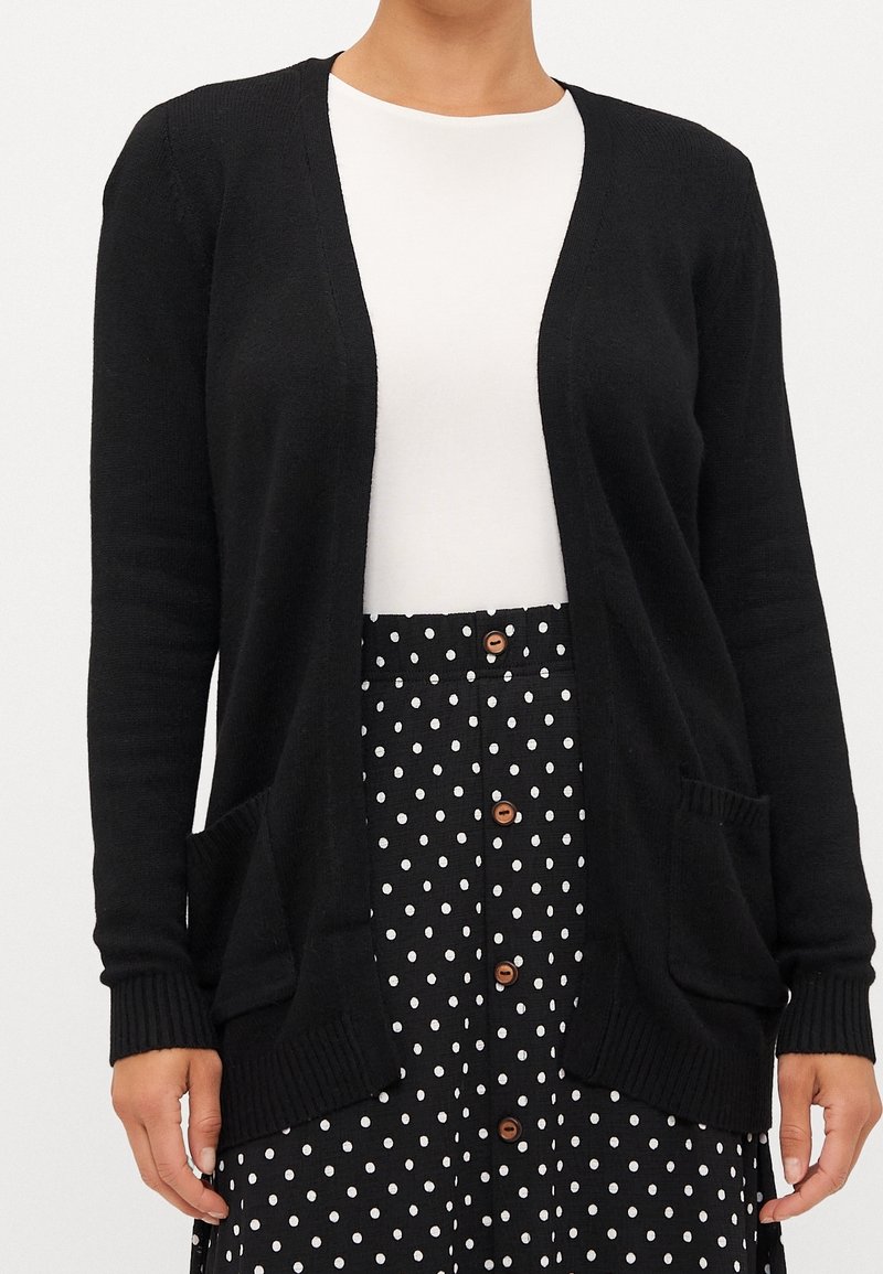 Femme portant un cardigan noir ouvert devant, un haut blanc et une jupe noire à pois boutonnée, montrant le buste et les mains.