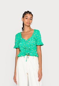 Top floral verde de manga curta com frente franzida e detalhe de cordão na barra. Apresenta um tecido texturizado e mangas largas.