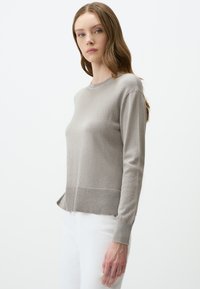 Pull gris clair à manches longues avec un col rond, tissu texturé, bord à côtes discret et silhouette ajustée, associé à un pantalon blanc.