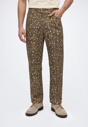 Un mannequin porte un pantalon brun olive parsemé de taches crème, une chemise blanche et des chaussures beiges ; une main repose dans une poche.