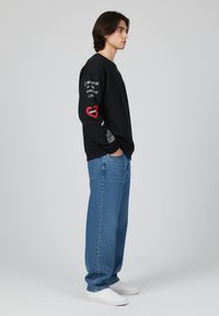 Felpa nera con testo grafico e cuore rosso sul manica; abbinata a jeans blu larghi e scarpe da ginnastica bianche. Stile semplice e casual.