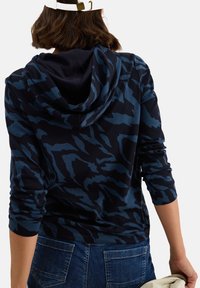 Vrouw in een blauw en zwart gemarmerde hoodie en denim jeans, houdt een licht jasje vast, op een witte achtergrond, gezien van achteren.