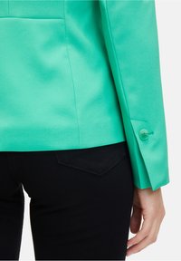 Tyrkysový blazer s hladkou texturou, s jediným knoflíkem na manžetě, nošený přes černé kalhoty s detaily kapsy.