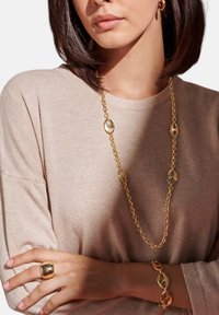 Schmuck aus Gold umfasst eine lange Kettenhalskette mit ovalen Gliedern, ein passendes Armband und einen auffälligen Ring. Das Model trägt einen beigen Pullover.