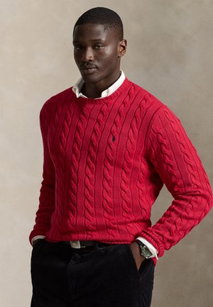Homme portant un pull en maille torsadée rouge par-dessus une chemise blanche à col, pantalon noir et une montre-bracelet, debout les mains dans les poches.