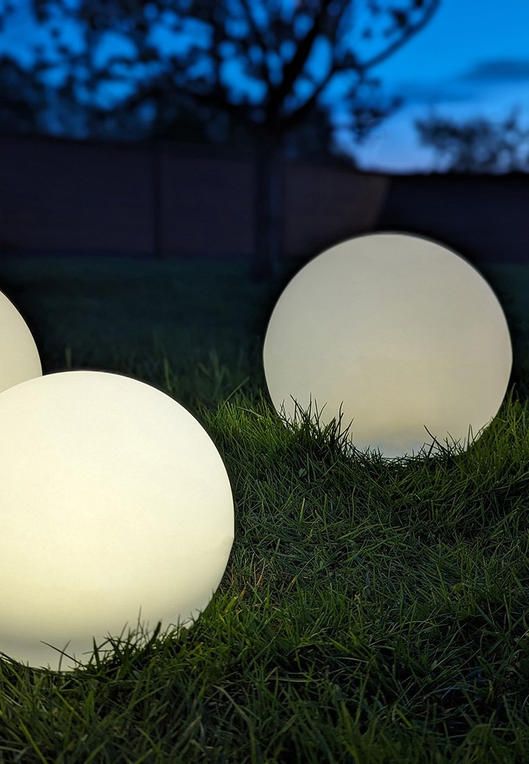 Tre grandi lampade sferiche bianche sull'erba, che brillano delicatamente al crepuscolo, con una finitura opaca liscia e senza componenti visibili.