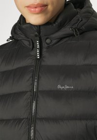 Chaqueta acolchada negra con cuello alto, cierre de cremallera y logo de "Pepe Jeans". Presenta una textura suave y un patrón de acolchado horizontal.