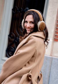 Femme en manteau beige et écouteurs marron souriant par-dessus son épaule, se tenant à l'extérieur près d'une porte fermée avec un décor en fer forgé.