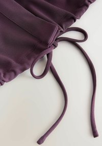 Fialová látka s nabráním a dvěma dlouhými, tenkými pásky. Materiál působí hladce, má měkkou texturu a konzistentní barvu po celé ploše.
