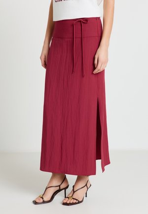 Femme portant une longue jupe rouge avec une fente devant et une taille à cordon, associée à des sandales à talons noires à brides.